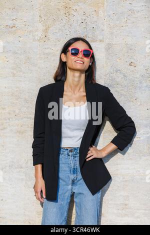 Frau in Sonnenbrille und lässigem Outfit lächelt, während sie an einer Steinmauer steht. Spanien Stockfoto