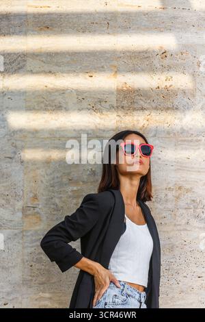 Frau in Sonnenbrille und lässigem Outfit posiert vor einer strukturierten Steinmauer. Spanien Stockfoto