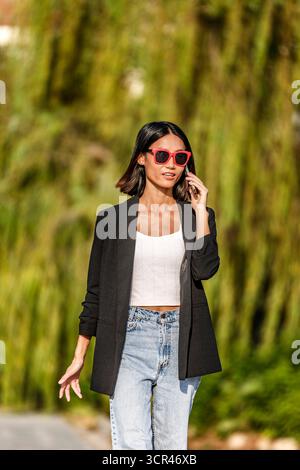 Frau in schwarzem Blazer und roter Sonnenbrille läuft, während sie draußen telefoniert. Spanien Stockfoto