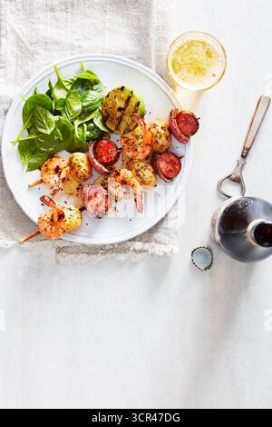 Gegrillte Garnelen- und Gemüsespieße auf einem weißen Teller mit frischem Spinat und einer Flasche Bier. Stockfoto