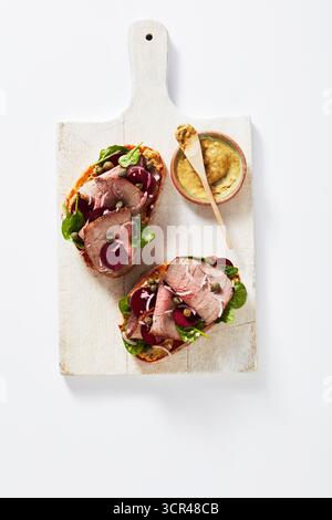 Zwei offene Roastbeef-Sandwiches mit Gemüse auf einem weißen Schneidebrett, mit Senf an der Seite. Stockfoto