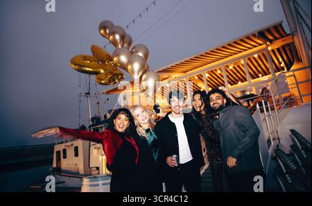 Eine energiegeladene Gruppe von Freunden, die bei Nacht an Bord einer Yacht eine lebhafte Feier genießen. Goldene Ballons verleihen diesem Mem eine festliche und fröhliche Urlaubsstimmung Stockfoto