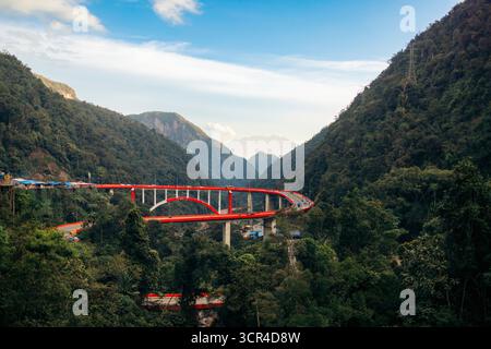 Eine leuchtende rote Brücke schlängelt sich durch üppige grüne Berge unter einem klaren blauen Himmel. Kelok 9 oder die 9 Haarnadeln, Harau, West Sumatra, Indonesien Stockfoto