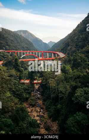 Die rote Brücke schlängelt sich durch üppige grüne Berge unter einem klaren blauen Himmel. Kelok 9 oder die 9 Haarnadeln, Harau, West Sumatra, Indonesien Stockfoto