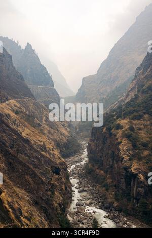 Majestätisches Bergtal mit einem sich windenden Fluss und steilen, zerklüfteten Klippen unter einem nebeligen Himmel. Straße F36 von Annapurna in der Provinz Gandaki, Nepal Stockfoto
