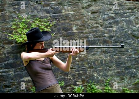 Frau mit schwarzem Hut, die draußen mit einem Gewehr auf eine von Grün umgebene Zielreichweite zielt. Stockfoto