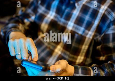 Mann in lässigem kariertem Hemd, der das Smartphone mit beiden Händen hält, während er drinnen unter künstlichem Licht den Bildschirm durchblättert Stockfoto