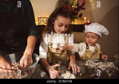 Eine Mutter und zwei Kinder backen Lebkuchen und Formen Teig in einer festlichen, warm beleuchteten Küche. Schürzen auf, Mehl staubt den Tisch, während die Lichter leuchten Stockfoto