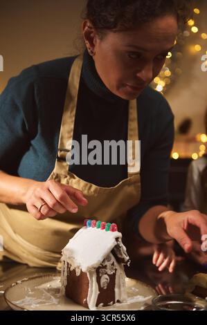 Eine Frau in einer beigefarbenen Schürze ziert ein Lebkuchenhaus und setzt Süßigkeiten in einem warmen, festlichen Zuhause. Die Urlaubsszene fängt Backkunst, Familienstimmung, Stockfoto