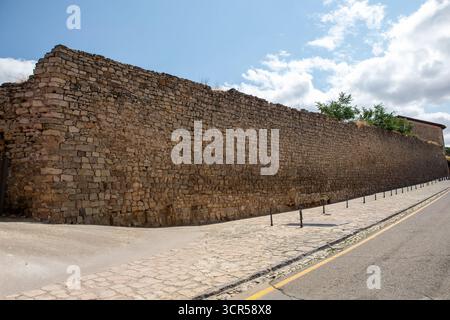Die historische Steinmauer erstreckt sich entlang einer kopfsteingepflasterten Straße und zeigt alte Handwerkskunst unter einem sonnigen Himmel mit Wolken. Das nahe gelegene Grün trägt zur Landschaft bei. Stockfoto