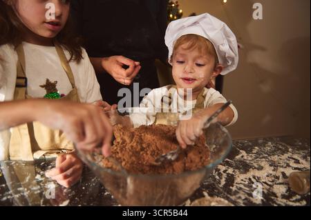 Ein fröhliches Kind mit Kochmütze mischt Lebkuchenteig in einer Glasschale auf einem überdachten Küchentisch, geführt von einem Erwachsenen. Die Szene fängt Fam ein Stockfoto