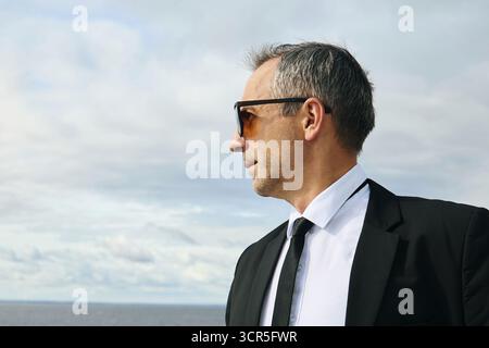 Kaukasischer reifer Mann mit Sonnenbrille, der draußen steht und mit ernstem Ausdruck von der Kamera wegschaut, kurzen Haaren sichtbar, Himmel und Wasser im Hintergrund Stockfoto
