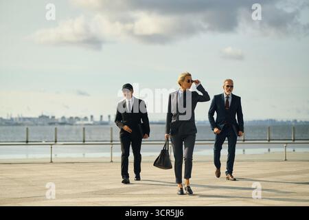 Kaukasische Geschäftsfrau mittleren Alters in Business-Anzug und Sonnenbrille, die selbstbewusst im Freien mit zwei männlichen Kollegen in Anzügen hinter der Nähe des Ufers im urbanen Hintergrund läuft Stockfoto