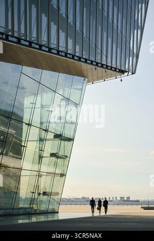 Drei junge Geschäftsleute laufen zusammen vor einem hohen Glasgebäude, einer entfernten Skyline der Stadt, die im Hintergrund sichtbar ist, und die Gruppe bewegt sich am sonnigen Tag entlang des Ufers Stockfoto