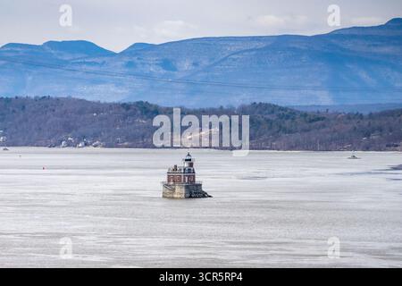 Aus der Vogelperspektive auf den ikonischen Leuchtturm Hudson-Athens, der widerstandsfähig inmitten des kalten Wassers steht, mit einer Kulisse aus trüben Bergen und heiterem Himmel, Huds Stockfoto