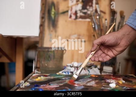 Künstler, der im Studio arbeitet. Männliche Hand hält Pinsel und mischt Ölfarben auf bunter, unordentlicher Palette, Nahaufnahme Stockfoto