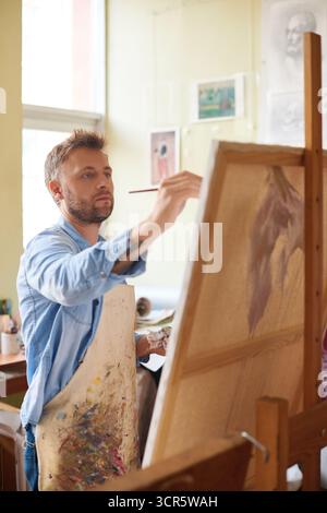 Lackierprozess. Bärtiger Künstler in Denim-Hemd und gebeizter Schürze, der vor der Staffelei steht und mit Begeisterung ein Bild malt Stockfoto