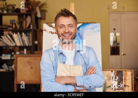 Erfolgreiches Kunstgeschäft. Porträt eines bärtigen Malers, der mit gefalteten Armen gegen seine Schöpfung steht und glücklich in die Kamera lächelt Stockfoto
