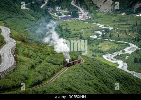 Aus der Vogelperspektive einer alten Dampfeisenbahn, die durch grüne Hügel entlang einer gewundenen Straße schlängelt, mit dem Dorf Obergoms im Tal, Ober Stockfoto