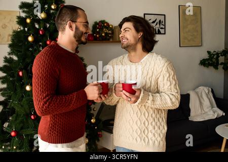 Zwei liebevolle Männer teilen fröhliche Momente zusammen und halten rote Tassen in festlicher Atmosphäre. Stockfoto