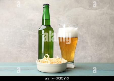 Getrocknete Tintenfischspäne und Bier auf hellblauem Holztisch vor grauem Hintergrund, Nahaufnahme Stockfoto