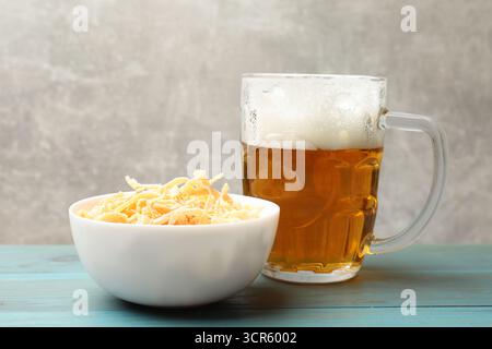 Getrocknete Tintenfischspäne und Glasbecher Bier auf hellblauem Holztisch vor grauem Hintergrund, Nahaufnahme Stockfoto