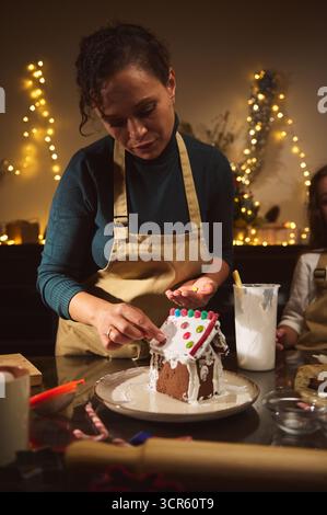 Eine Frau in einer beigefarbenen Schürze ziert ein winziges Lebkuchenhaus mit Glasur und bunten Süßigkeiten in einer warm beleuchteten, festlichen Küche. Stockfoto