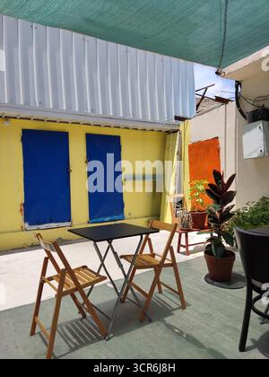 Eine lebendige und eklektische Terrassenszene im Freien mit einem kleinen schwarzen Metall-Bistrotisch mit zwei hellen Holzfaltstühlen, die in einem kräftigen Farbton gehalten werden Stockfoto