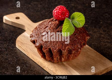 Dieses Nahbild zeigt einen köstlichen Muffin, der wunderschön mit frischen Himbeeren dekoriert ist und perfekt auf einem rustikalen Holzschneidebrett angeordnet ist. Stockfoto