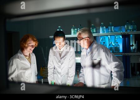 Lächelnde Medizinstudierende mit einem erfahrenen Mentor, der im Labor an ihrem Projekt arbeitet. Sie stehen am Tisch mit Reagenzgläsern mit bunten Flüssigkeiten Stockfoto