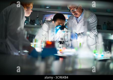 Multiethnische Gruppe von Chemikern, die im modernen Labor forschen. Hübsche junge Frau in Schutzmaske, die durch das Mikroskop schaut, während ihre Kollegen in der Nähe stehen Stockfoto