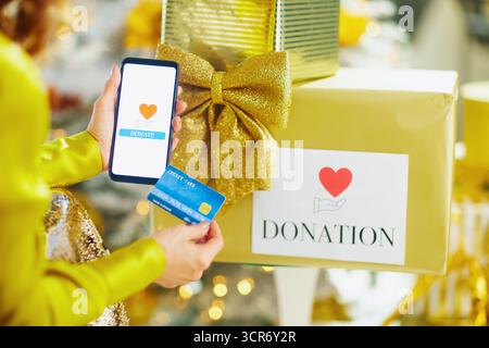 Eine Nahaufnahme zeigt Hände, die das Smartphone halten, und zeigt Spendenapp und Kreditkarte über einer goldenen Geschenkbox. Der festliche Hintergrund von verschwommenen Lichtern wird hervorgehoben Stockfoto