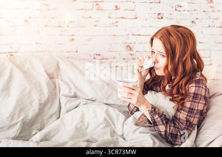 Eine Frau, die krank auf einem Sofa sitzt, mit einer Teetasse in der Hand und ihr die Nase bläst Stockfoto