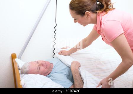 Senior im Pflegeheim im Bett, Krankenschwester deckt ihn ab Stockfoto