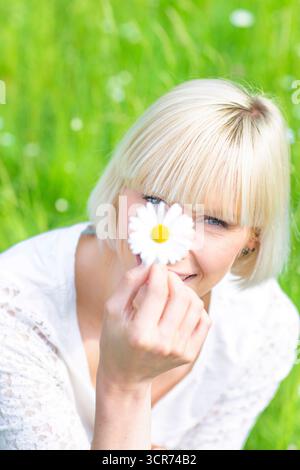 Glückliche Frau, die auf einer Wiese sitzt und ein Gänseblümchen in der Hand hält und in die Kamera schaut. Stockfoto