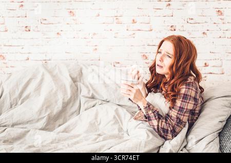 Eine Frau, die krank auf einem Sofa sitzt, mit einer Teetasse in der Hand und ihr die Nase bläst Stockfoto