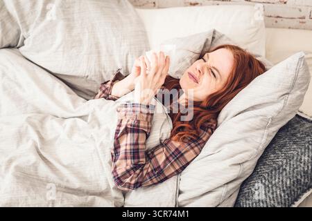 Eine Frau, die krank auf einem Sofa liegt und sich die Nase bläst Stockfoto