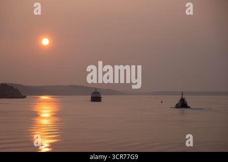 COBH, IRLAND – 15. August 2025: Ein Schiff des Irish Naval Service und ein Frachtschiff fahren bei Sonnenuntergang durch den Hafen von Cork Stockfoto