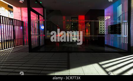 Gebäudeeingang mit Glas, das die umgebende Architektur reflektiert, und farbigen Lichtern. Eine Mischung aus Licht und Schatten mit einem Kühlergrill und runden Balken. Stockfoto