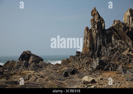 Malerische Aussicht auf scharfe massive Felsen und Steinformationen, die sich am Strand erheben, gegen abstürzende Wellen auf dem Meer oder der Küste, vor klarem blauem Himmel Stockfoto