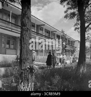 Ein Blick auf ein zweistöckiges Sanatorium oder Ferienhaus mit einem großen Propagandaplakat mit einem heldenhaften Arbeiter an der Wand im Slavjansk Resort (Slavkurort), wo Gruppen von Urlaubern das Parkgelände, ukrainische SSR, zwischen den 1970er und 1980er Jahren, genießen Dieses stimmungsvolle Schwarzweiß-Vintage-Foto fängt die typische Architektur und das tägliche Leben eines sowjetischen Resorts ein, in dem die Freizeit von staatlicher Ideologie umrahmt wurde, was den einfachen, staatlich bereitgestellten Urlaub in der friedlichen Donbass-Region vor dem Krieg symbolisiert. Stockfoto