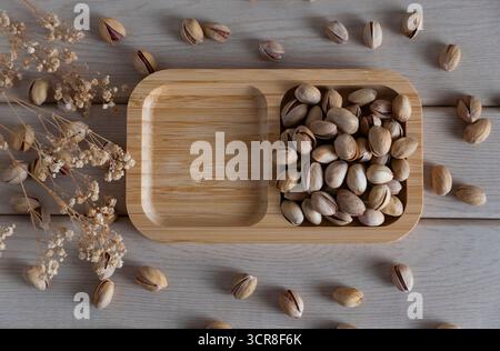 Blick von oben auf rustikale Pistazien auf einem Holztablett, verstreut auf einer hellen Holzoberfläche, die natürliche Texturen und Erdtöne hervorhebt Stockfoto