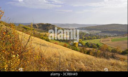 Ländliche Landschaft im Herbst. Blick auf das Dorf in den Bergen, Siebenbürgen. Sanfte Hügel, die mit goldenem Herbstgras bedeckt sind, führen zu einer malerischen Landschaft. Stockfoto