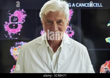 Porträt eines selbstbewussten älteren kaukasischen männlichen Arztes, der vor der wissenschaftlichen Anzeige lächelt und steht, Bilder der Krebszellforschung zeigt, Laborkittel trägt und medizinische Daten auf der Konferenz präsentiert Stockfoto