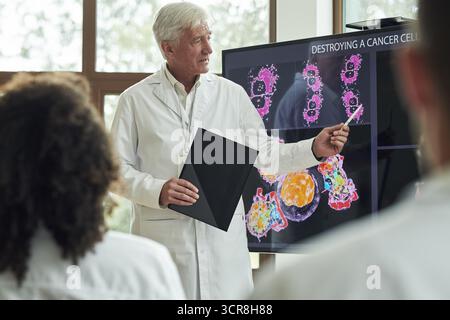 Senior-kaukasischer männlicher Professor erklärt der Gruppe von jungen Studenten in der medizinischen Ausbildung den Prozess der Krebszellzerstörung, hält Ordner und zeigt auf wissenschaftliche Bilder auf dem Bildschirm Stockfoto