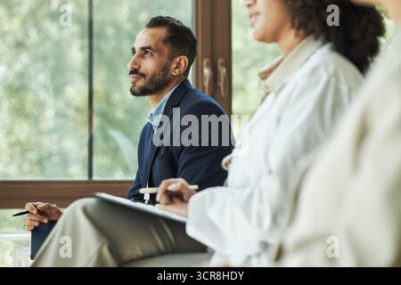 Junger polyethnischer Mann, der aufmerksam sitzt während eines Treffens mit weiblichen Kollegen, macht Notizen im Vordergrund, im großen Fenster und im Hintergrund, bei Konferenzen oder Seminaren sichtbares Grün Stockfoto
