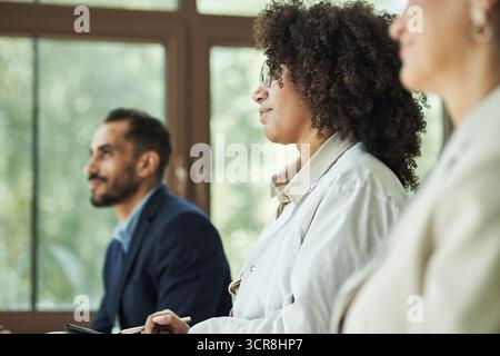 Junge Schwarze Frau im weißen Labormantel, die während eines Healthcare Business Meetings unter einer Gruppe von Fachleuten sitzt und aufmerksam mit verschiedenen Kollegen im Konferenzumfeld zuhört Stockfoto