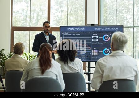 Junger Mann im Anzug aus dem Nahen Osten, der medizinische Daten über die Prävention von Diabetes und medizinische Nebenwirkungen vor einer Gruppe von multiethnischen Ärzten älteren und mittleren Alters im Konferenzraum vorstellt Stockfoto