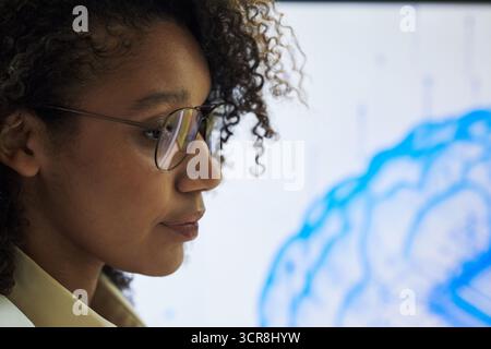 Junge Schwarze Frau mit Brille, die auf einer medizinischen Konferenz steht und sich auf die digitale Gehirnillustration konzentriert, die auf dem Bildschirm angezeigt wird, um das wissenschaftliche Forschungskonzept zu demonstrieren Stockfoto