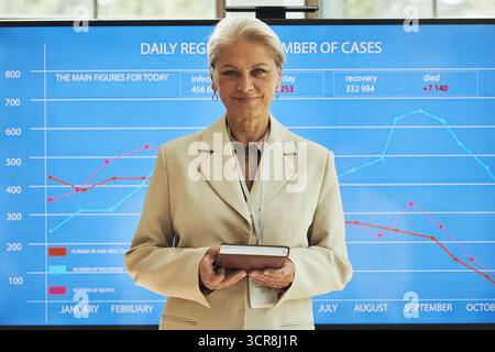 Porträt einer älteren kaukasischen Frau, die vor einer großen digitalen Karte steht, die ein Notizbuch hält, selbstbewusst lächelnd und statistische Daten über die täglich registrierte Anzahl medizinischer Fälle auf der Konferenz präsentiert Stockfoto